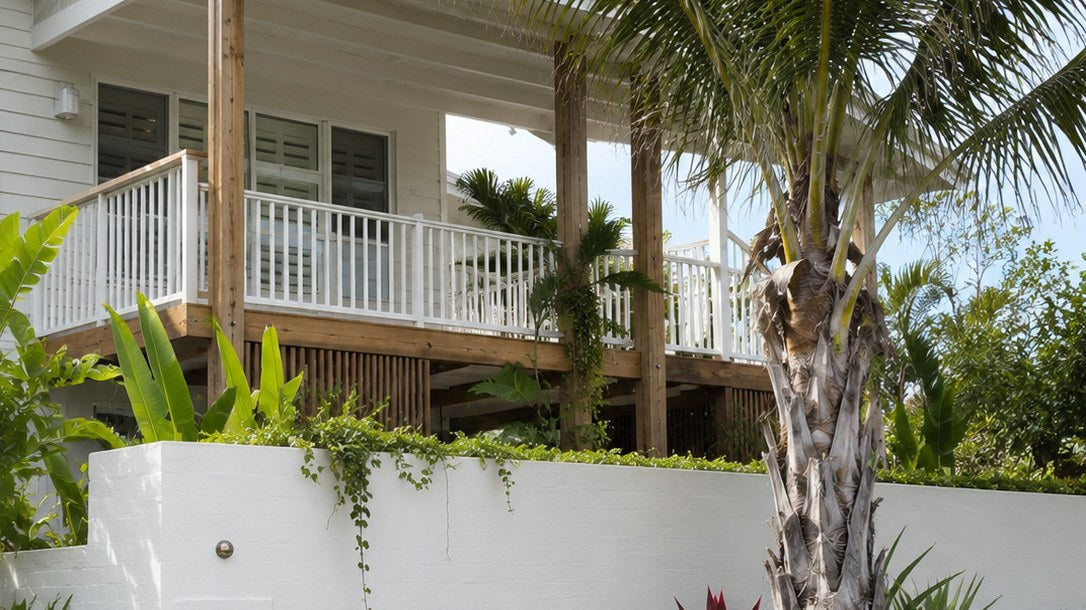 Corner garden of a white weatherboard coastal home with an elevated timber-post verandah, a mature pinnate palm rising past a white brick fence, sandstone boulders, agave, red cordyline and tropical understorey over lawn.